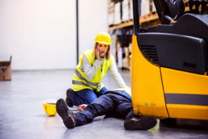 woman wearing yellow vest and hard hat calling for help on a phone next to a person lying down on the floor next to a forklift after an accident